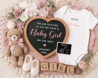Könnte beinhalten: Ein herzförmiges Schild aus Schiefer mit dem Text "We are excited to say we have a Baby Girl on the way! January 2026". Neben dem Schild liegt ein weißer Strampler mit dem Text "Baby Wilson". Ein Teddybär, ein Paar Babyschuhe und Holzklötze, die "GIRL" buchstabieren, sind ebenfalls auf dem Bild zu sehen.