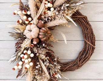 May include: A circular wreath constructed from brown twisted twigs, adorned on one side with a collection of dried grasses, metallic silver leaves, white berries, and two small pumpkins in shades of beige and bronze-brown.