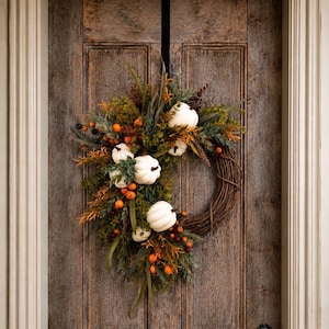 May include: A circular wreath constructed from brown woven twigs, adorned with white pumpkins, green foliage, and small orange and dark berries. The arrangement is clustered along the left side and top of the wreath, leaving a portion of the twigs exposed.