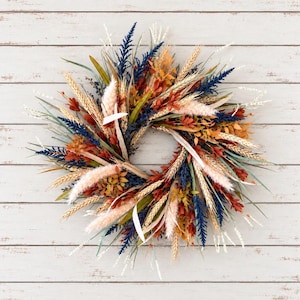May include: A circular wreath composed of dried botanical elements including navy blue sprigs, golden wheat stalks, rust-colored foliage, and soft cream-colored pampas grass plumes arranged in a dense, textured, and multi-layered radial pattern.