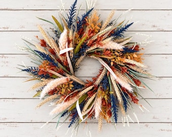 May include: A circular wreath composed of dried botanical elements including navy blue sprigs, golden wheat stalks, rust-colored foliage, and soft cream-colored pampas grass plumes arranged in a dense, textured, and multi-layered radial pattern.