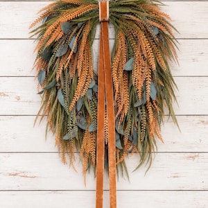 May include: A teardrop-shaped wreath featuring a mix of green and orange foliage, including fern-like sprigs and eucalyptus leaves. The arrangement is tied at the top with a long, smooth, copper-colored velvet ribbon that hangs down.