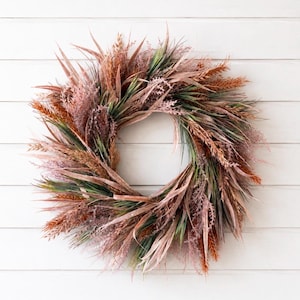 May include: A circular wreath composed of dried-style grasses and foliage in shades of muted rust, dusty pink, and deep forest green. The arrangement features long, slender blades and feathery textures radiating outward from a central open ring.