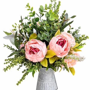 May include: A floral arrangement featuring large pink peonies with dark red centers, surrounded by green foliage and small white flowers. The bouquet is in a galvanized metal vase, creating a rustic aesthetic.