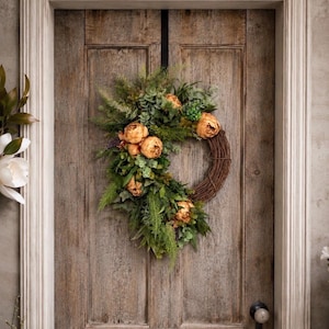 May include: A decorative wreath featuring a circular brown twig base adorned with a cluster of muted orange flowers, various green foliage, and ferns. The wreath is suspended by a black metal hanger on a weathered wooden door with a round knob.