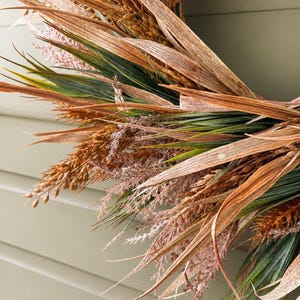 May include: A circular wreath composed of dried wheat stalks, long brown grass blades, and delicate feathery pinkish-tan plumes. The arrangement features a mix of textures and earthy tones, including deep green foliage interspersed with golden-brown grains.