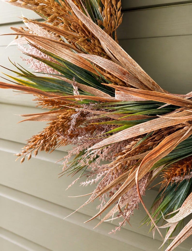 May include: A circular wreath composed of dried wheat stalks, long brown grass blades, and delicate feathery pinkish-tan plumes. The arrangement features a mix of textures and earthy tones, including deep green foliage interspersed with golden-brown grains.