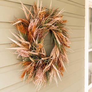 May include: A circular wreath composed of dried grasses, wheat stalks, and feathery plumes in shades of muted tan, light brown, sage green, and soft dusty pink. The arrangement features varied textures of pointed leaves and grain-like botanical elements.
