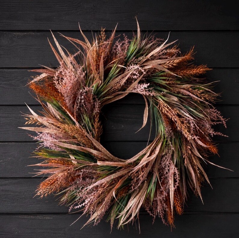 May include: A circular wreath composed of dried-style foliage in shades of muted copper, rust, dusty pink, and deep forest green. The arrangement features long, slender, pointed leaves mixed with feathery, textured grass-like stems and stalks.