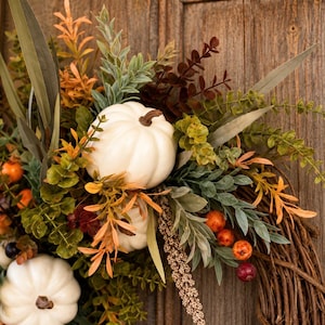 May include: A decorative wreath featuring white pumpkins, green and orange foliage, and small berries arranged on a brown twig base. The arrangement includes long, pointed leaves, textured sprigs, and round orange and dark red berries.