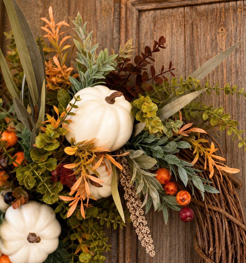 May include: A decorative wreath featuring white pumpkins, green and orange foliage, and small berries arranged on a brown twig base. The arrangement includes long, pointed leaves, textured sprigs, and round orange and dark red berries.