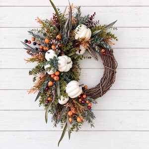 May include: A circular wreath constructed from brown woven twigs, adorned with white pumpkins, green eucalyptus leaves, and clusters of small orange and dark purple berries. The arrangement is asymmetrical, featuring foliage extending outward from the base.