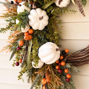 May include: A decorative wreath featuring white pumpkins with brown stems, interspersed with green eucalyptus leaves, orange berries, and dark purple berries. The arrangement is built upon a natural brown twig base, creating a seasonal display.