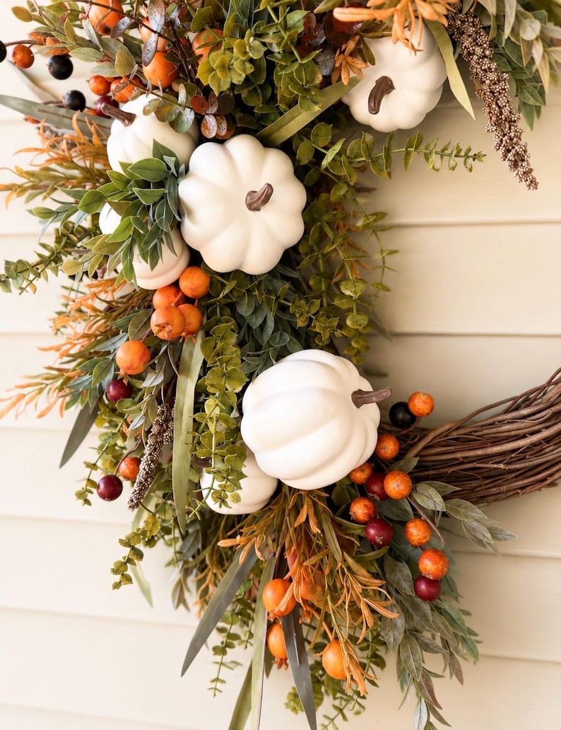May include: A decorative wreath featuring white pumpkins with brown stems, interspersed with green eucalyptus leaves, orange berries, and dark purple berries. The arrangement is built upon a natural brown twig base, creating a seasonal display.