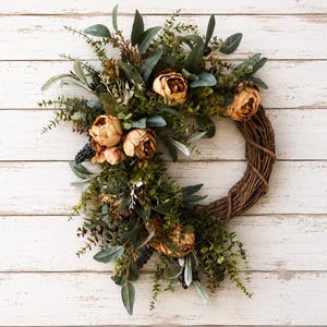 May include: A circular wreath constructed from a brown woven twig base, adorned with a dense arrangement of green eucalyptus leaves, small dark berries, and several large, muted orange-toned peony flowers clustered on one side.