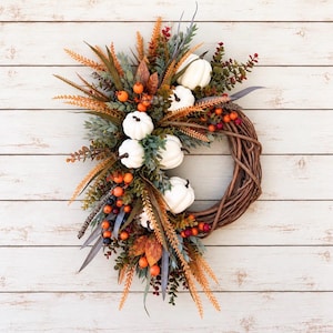 May include: A circular wreath constructed from brown woven twigs, adorned on one side with white artificial pumpkins, orange and dark blue berries, and various green and orange foliage including fern-like sprigs and leaves.