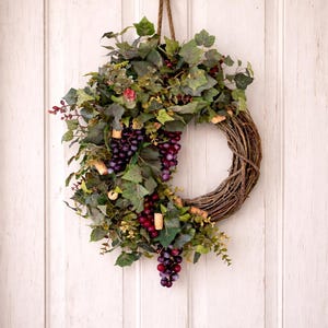 May include: A circular wreath constructed from brown twisted twigs, adorned with artificial green ivy leaves, clusters of deep purple grapes, and small light brown cylindrical wine corks, suspended by a thick jute rope from a black metal hook.