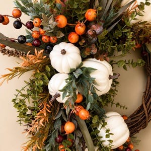 May include: A decorative wreath featuring white pumpkins, green eucalyptus leaves, and clusters of small orange and dark purple berries. The arrangement is set on a brown woven twig base, incorporating various autumn-toned foliage and stems.