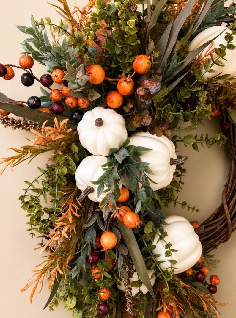 May include: A decorative wreath featuring white pumpkins, green eucalyptus leaves, and clusters of small orange and dark purple berries. The arrangement is set on a brown woven twig base, incorporating various autumn-toned foliage and stems.