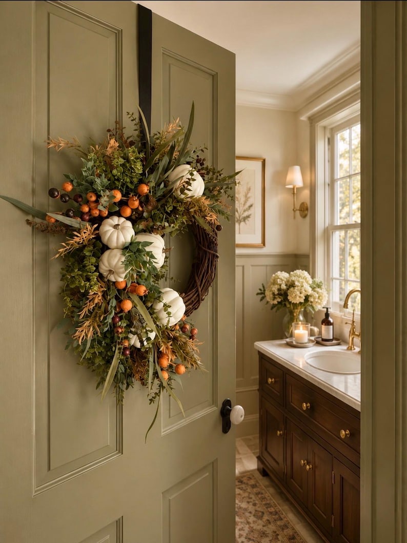 May include: A decorative wreath hanging on a sage green door, featuring a woven brown twig base adorned with white pumpkins, orange berries, and various green foliage, suspended by a black strap attached to the top of the door frame.