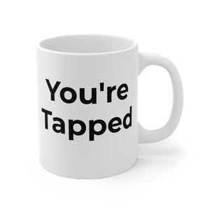 Könnte beinhalten: Ein weißer Keramikbecher mit dem schwarzen Text You're Tapped auf der Vorderseite. Der Becher hat einen zylindrischen Körper mit glatter Oberfläche und einen geschwungenen Henkel auf der rechten Seite. Das Design ist schlicht, klar und zentriert.
