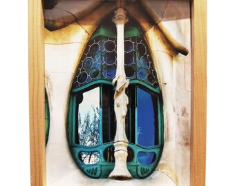 May include: A framed photograph of a unique, oval-shaped window with stained glass and a blue sky reflection. The window is set in a white, textured wall.