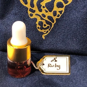 Op de afbeelding: Een klein glazen flesje met een gouden dop en een rode vloeistof erin. Het flesje is gelabeld met "Ruby" en staat op een blauw fluwelen oppervlak. Het label heeft ook een klein gouden symbool van een schaar.