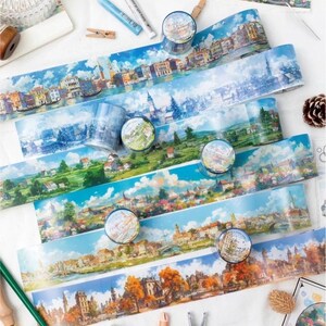 May include: A selection of colourful washi tapes featuring scenic cityscapes with blue skies and white clouds. The tapes are rolled up and arranged on a white surface with other craft supplies.