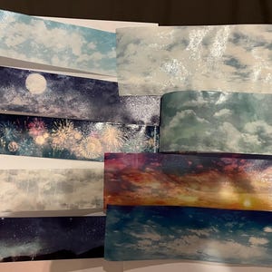 Peut inclure: Une collection de rubans adhésifs décoratifs washi avec des motifs de ciel et de nuages. Les rubans présentent diverses scènes, notamment un ciel nocturne avec une pleine lune, des feux d'artifice et un coucher de soleil sur l'océan. Les rubans sont dans des tons de bleu, blanc et rose.