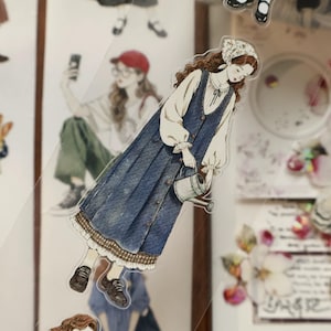 May include: A sticker featuring a watercolor illustration of a woman in a blue dress holding a watering can. The dress has a button-down front and a decorative trim. The woman wears a white blouse and a floral headscarf. The sticker is transparent.