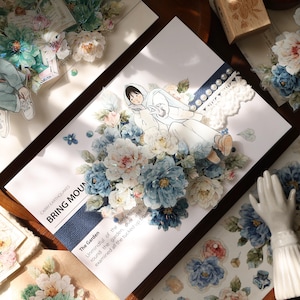 May include: A decorative paper with a floral design featuring blue and white peonies, a cartoon figure, and the text "BRING MOUN". The paper is embellished with lace and pearls, creating a vintage aesthetic.