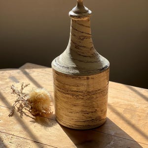 Peut inclure: Un vase en céramique haut et de couleur crème avec un motif brun en bois. Le vase a une base large et un col étroit avec un sommet arrondi.