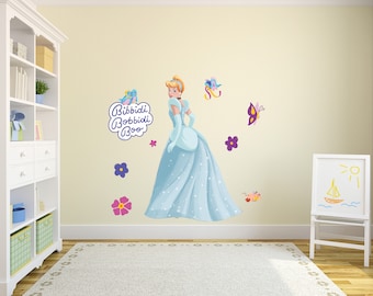 Op de afbeelding: Een lichtblauwe muursticker met Assepoester in een blauwe baljurk met een paarse vlinder en bloemen. De tekst "Bibbidi Bobbidi Boo" staat in een paarse wolk.