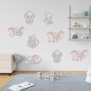 May include: A set of nine grey and pink Dumbo the Elephant wall decals. The decals are arranged on a white wall in a nursery setting.