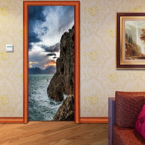 Puede incluir: Una calcomanía para puerta que presenta un paisaje escénico con un acantilado rocoso que domina un mar tormentoso. El cielo es una mezcla dramática de nubes oscuras y una puesta de sol naranja brillante.
