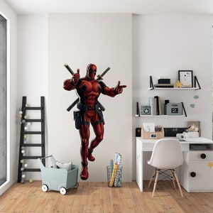 Könnte beinhalten: Ein Wandaufkleber mit dem roten und schwarzen Deadpool-Charakter mit zwei Schwertern hinter ihm. Er zeigt mit einer Hand den Daumen nach oben und zeigt mit der anderen. Der Aufkleber befindet sich an einer weißen Wand in einem Raum mit einem Schreibtisch, einem Stuhl und Regalen.