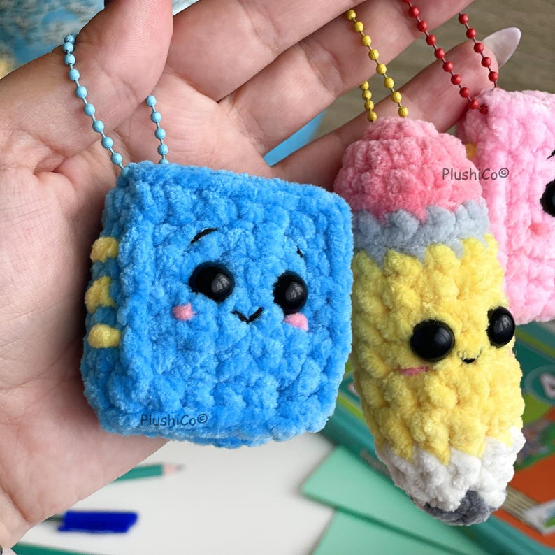 May include: Close-up of three handmade crochet keychains. The first is a blue cube with a smiling face and yellow accents. The second is a yellow pencil with a pink eraser and gray tip. The third is a pink keychain with a smiling face.