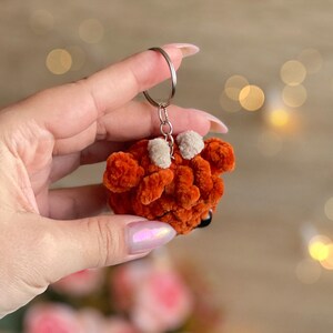 May include: A handmade orange plush keychain with a silver keyring. The plush has two small white accents and a black nose. The keychain is held in a hand against a blurred background.