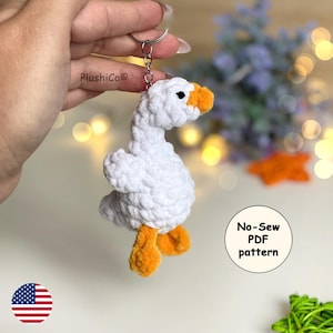 May include: A white crochet goose keychain with orange beak and feet. The keychain is attached to a silver ring. The image includes the text "No-Sew PDF pattern". The goose is held in a person's hand.