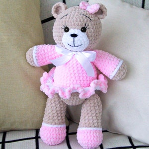 May include: A crocheted teddy bear wearing a pink dress with a white bow. The bear has brown fur and black eyes.