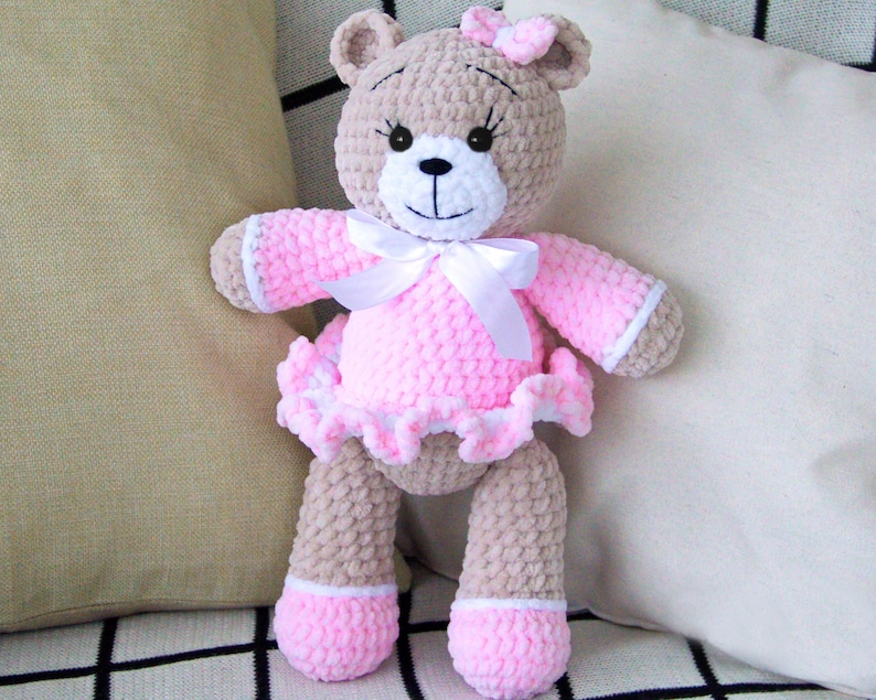 May include: A crocheted teddy bear wearing a pink dress with a white bow. The bear has brown fur and black eyes.