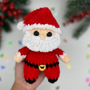 May include: A small, handmade crochet Santa Claus doll made of soft red, white, and black yarn. The figure features a red hat with a white pom-pom, a thick white beard, black safety eyes, and a black belt with a yellow buckle.