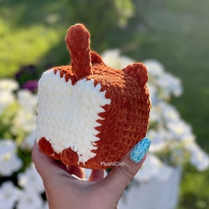 May include: A crocheted fox plush toy with a white belly and orange fur. The toy is being held by a hand with blue nail polish.