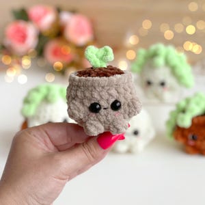 May include: A small, plush, crocheted plant in a tan pot with a brown top and a light green sprout. The pot features two black plastic eyes and small pink stitched cheeks. The texture is soft and fuzzy, crafted from yarn in a rounded shape.