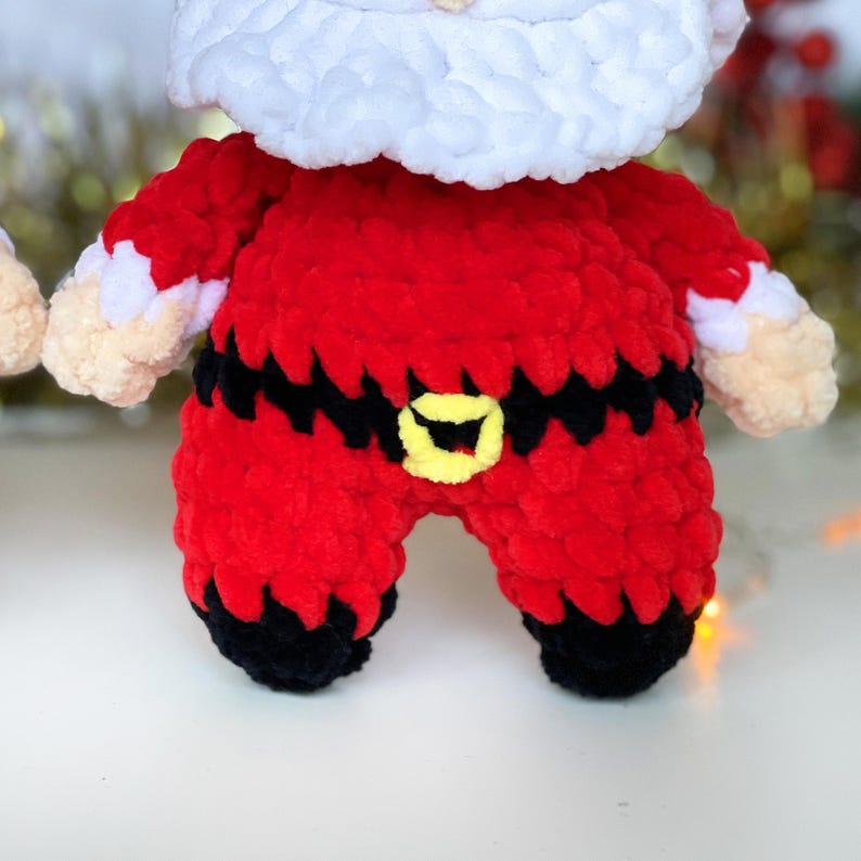 Puede incluir: Un mu&ntilde;eco de crochet de Pap&aacute; Noel con un traje rojo, un cintur&oacute;n negro y una hebilla amarilla. El mu&ntilde;eco tiene barba blanca y un gorro blanco.