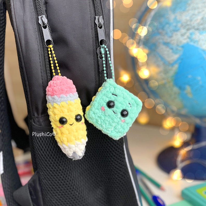 May include: Two crocheted backpack charms: a yellow pencil with pink eraser and a light green square. Both have smiling faces and are attached to a black backpack with zipper pulls. The pencil charm is approximately 3 inches long.