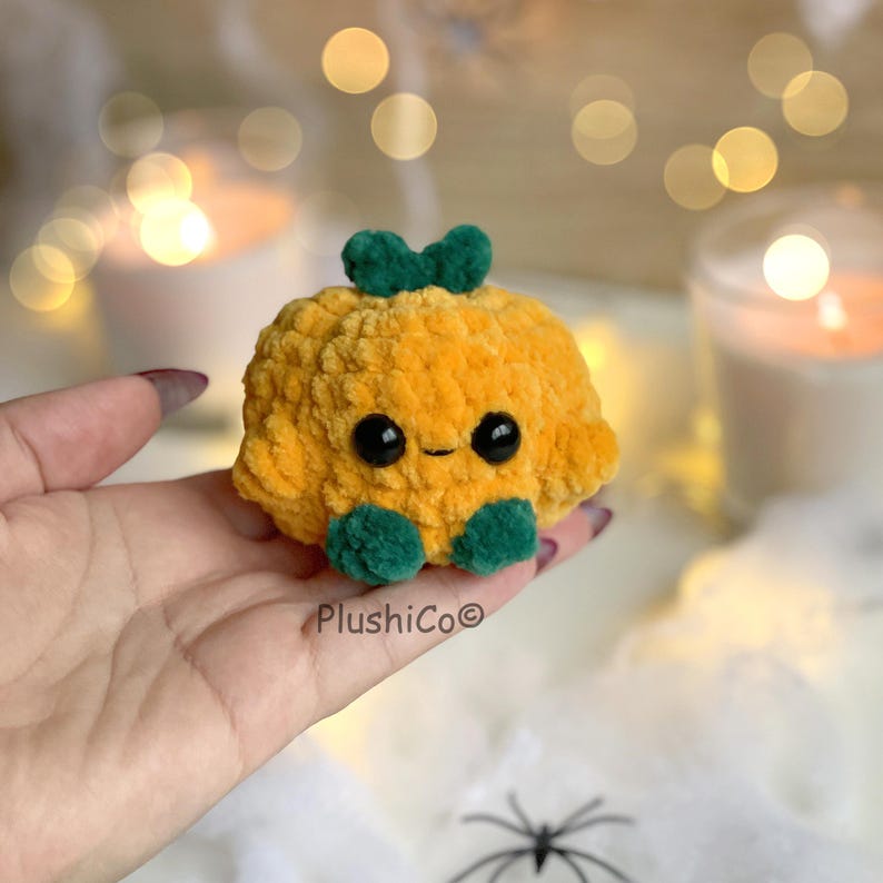 May include: A plush, orange pumpkin with a green stem and feet. The pumpkin has large black eyes and a small smile. The product is handmade and has the text "PlushiCo" on the bottom.