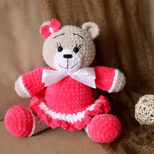 May include: A crocheted teddy bear wearing a pink dress with white trim and a white bow. The bear has brown eyes and a black nose.