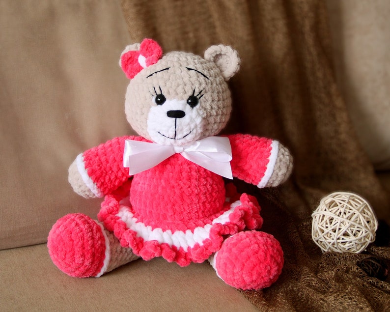 May include: A crocheted teddy bear wearing a pink dress with white trim and a white bow. The bear has brown eyes and a black nose.