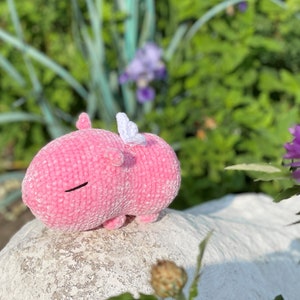 K&ouml;nnte beinhalten: Ein rosa geh&auml;keltes Stofftier mit wei&szlig;en Fl&uuml;geln. Das Tier liegt auf einem wei&szlig;en Stein mit gr&uuml;nen Pflanzen im Hintergrund.